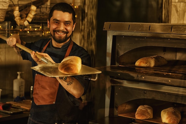 Lächelnder und schöner Bäcker mit Bart, der eine Schaufel mit frisch gebackenem Weißbrot hält Schöner Mann in Schürze, der in der Restaurantküche in der Nähe eines speziellen Konvektionsofens steht