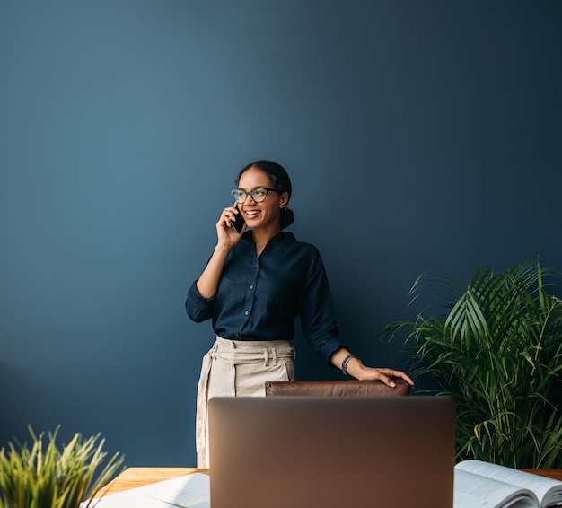 Lächelnder Unternehmer in formeller Kleidung, der zu Hause an einer blauen Wand steht und mit einem Mobiltelefon spricht