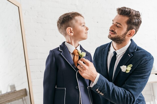 Lächelnder Vater und Sohn in Jacken mit Boutonnieres, die einander anschauen