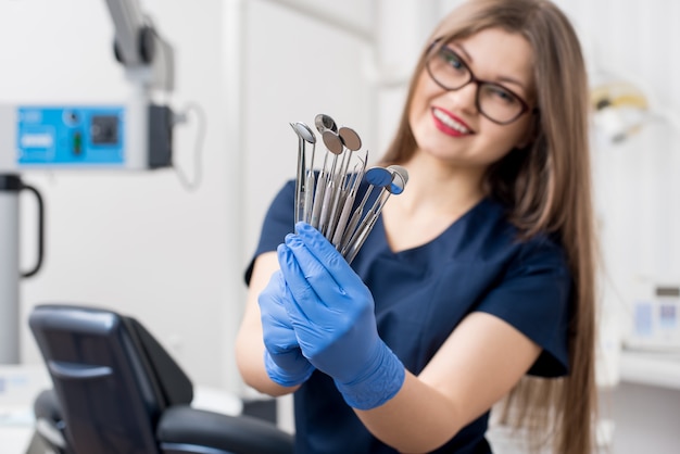 Lächelnder weiblicher Zahnarzt mit den blauen Handschuhen, die Werkzeuge - zahnmedizinische Spiegel und zahnmedizinische Sonden im zahnmedizinischen Büro halten
