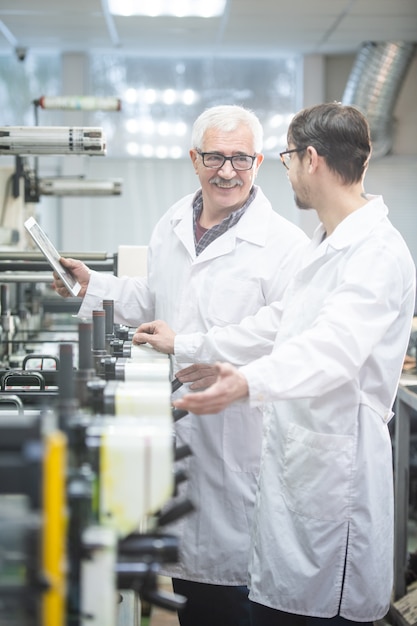 Lächelnder weißhaariger Arbeiter im Laborkittel unter Verwendung eines Tablets während der Analyse der Druckmaschinenproduktivität mit einem Kollegen im Fabrikladen