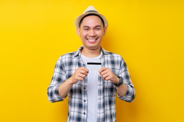 Lächelnder zufriedener asiatischer Ethnizität junger Mann der 20er Jahre mit lässiger Kleidung, Hut mit Kreditkarte, der selbstbewusst in die Kamera schaut, isoliert auf gelbem Hintergrund Studioporträt Menschen Lifestyle-Konzept