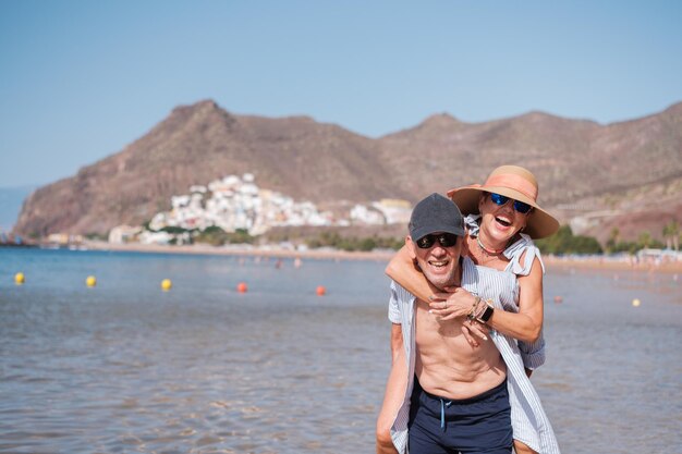 Lächelndes älteres Paar, das sie auf ihn klettert und am Strand Glück zeigt. Konzept, Glück, Ruhestand, Gesundheit