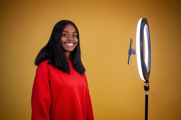 Lächelndes afrikanisch-amerikanisches Vlogger-Mädchen in einem roten Hoodie mit einer beleuchteten Ringlampe, die in die Kamera schaut