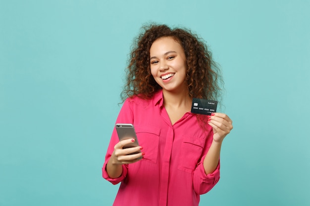 Lächelndes afrikanisches Mädchen in rosafarbener Freizeitkleidung mit Handy, das die Kreditkarte einzeln auf blauem türkisfarbenem Hintergrund im Studio hält. Menschen aufrichtige Emotionen Lifestyle-Konzept. Kopieren Sie Platz.