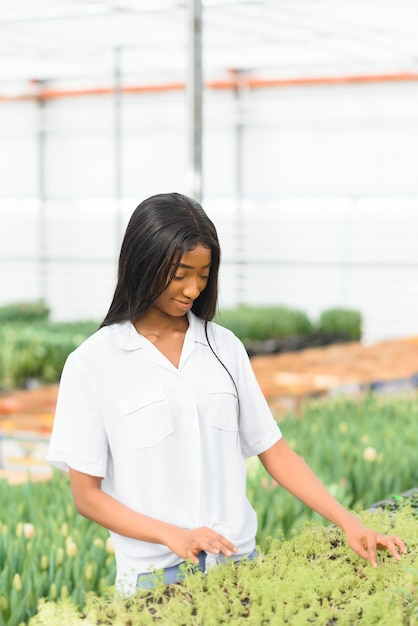 Lächelndes Afroamerikanermädchen prüft Blumen im Gewächshaus mit Tablette in ihren Händen