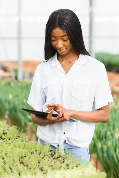 Lächelndes Afroamerikanermädchen prüft Blumen im Gewächshaus mit Tablette in ihren Händen