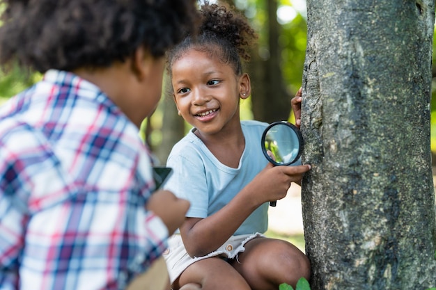 Lächelndes afroamerikanisches kleines Mädchen mit Freunden hält Lupe, um auf Baum im Park zu erkunden