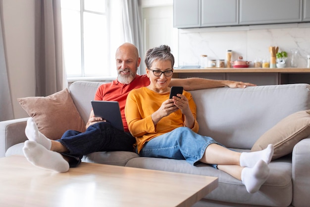 Lächelndes altes Paar ruht auf dem Sofa mit digitalem Tablet und Mobiltelefon