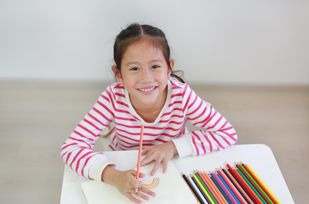 Lächelndes asiatisches kleines Mädchen, das zu Hause zeichnet und die Kamera nach oben schaut