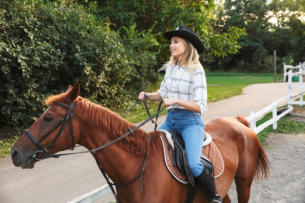 Lächelndes attraktives junges blondes Mädchen, das ein Pferd am Pferdehof reitet