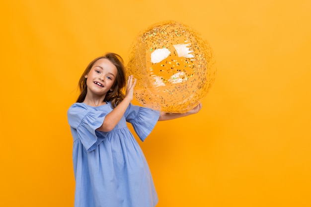 Lächelndes attraktives Mädchen in einem blauen Kleid hält einen funkelnden transparenten Ball auf Gelb mit Kopienraum