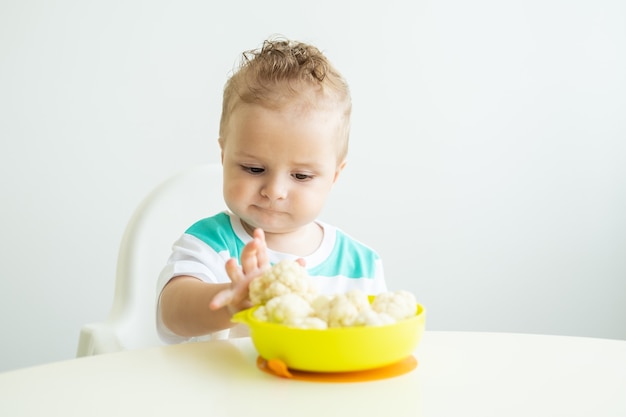 Lächelndes Baby, das in einem Kinderstuhl sitzt und Blumenkohl auf weißer Küche isst