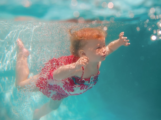 Lächelndes Baby in süßem, modernem Kleid, das unter Wasser im blauen Swimmingpool taucht