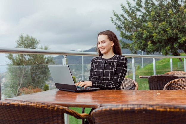 Lächelndes Bloggermädchen arbeitet online am Laptop, indem es auf dem Hintergrund der Naturansicht sitzt