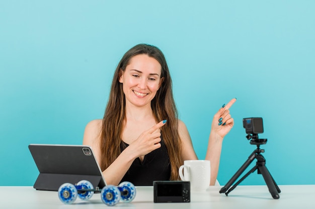 Lächelndes Bloggermädchen schaut auf ihre kleine Kamera, indem sie mit dem Zeigefinger auf blauem Hintergrund nach rechts zeigt