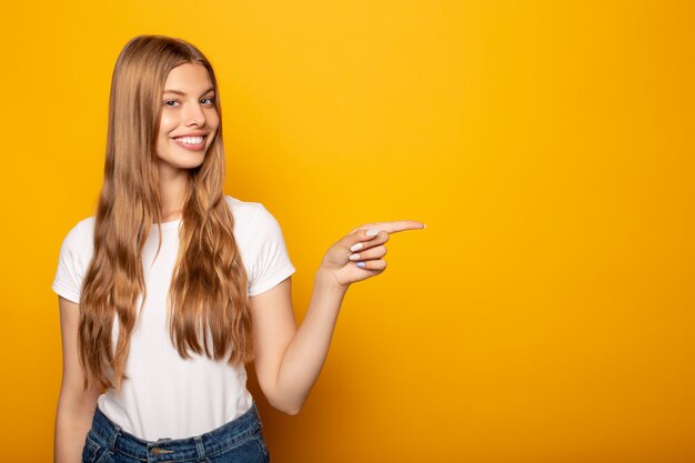 lächelndes blondes Mädchen, das mit Finger beiseite zeigt, lokalisiert auf Gelb