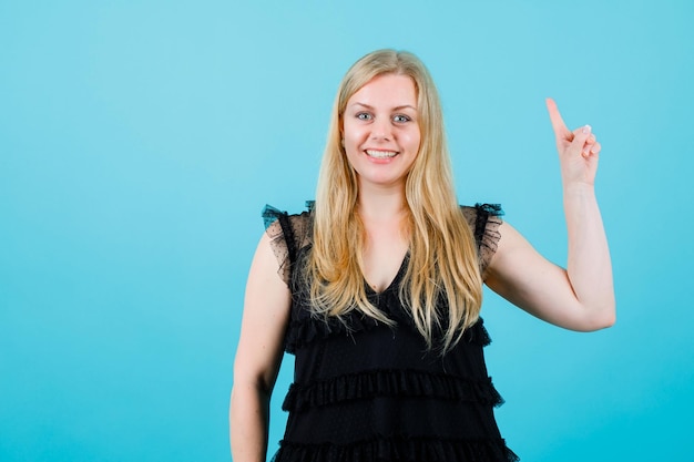 Lächelndes blondes Mädchen schaut in die Kamera, indem es mit dem Zeigefinger auf blauem Hintergrund nach oben zeigt