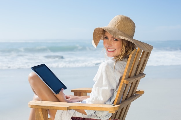 Lächelndes blondes Sitzen auf hölzernem Klappstuhl durch das Meer unter Verwendung der Tablette