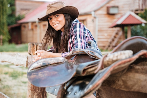 Lächelndes brünettes Cowgirl der jungen Frau, das sich auf Zaun im Dorf stützt