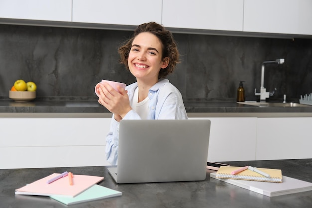 Lächelndes brünettes Mädchen sitzt mit Laptop in der Küche, trinkt Kaffee und arbeitet von zu Hause aus