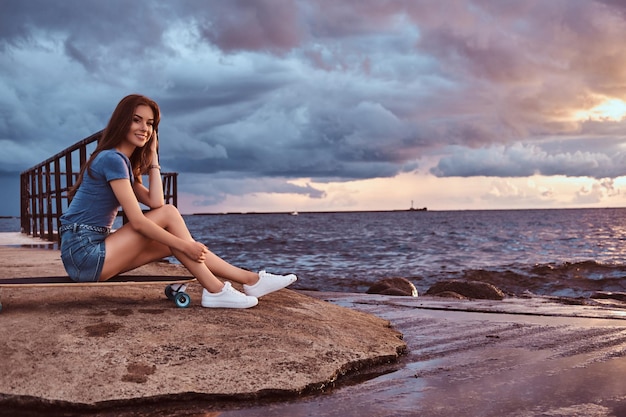 Lächelndes charmantes Mädchen, das auf einem Skateboard am Strand sitzt, genießt während des Sonnenuntergangs erstaunliches dunkles bewölktes Wetter.