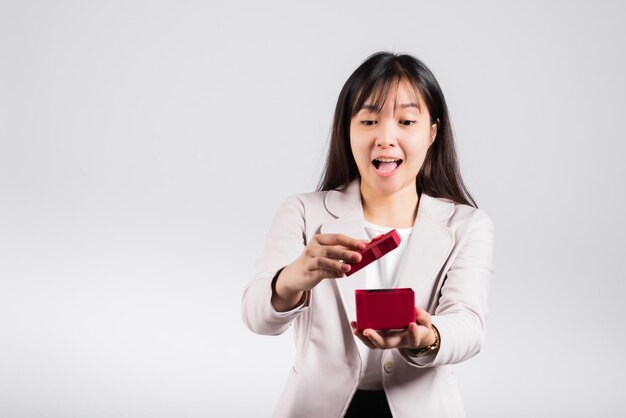 Lächelndes Frauenvertrauen, das eine rote Geschenkbox auf lokalisiertem weißem Hintergrund der Palme öffnet, Überraschungsgeburtstag oder Weihnachtstag und Valentinsgruß, asiatisches glückliches Porträt schöne junge weibliche Atelieraufnahme, Kopienraum