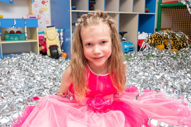 Lächelndes, fröhliches Mädchen in einem rosa Kleid in einem riesigen Haufen Silberfolienkonfetti im Kinderzimmer Ein Mädchen in funkelnden Pailletten