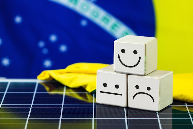 Lächelndes Gesicht und trauriges Gesicht in Holzblockwürfel und Brasilien-Flagge auf Photovoltaik-Solarpanel Positives Mindset-Konzept für saubere Energie