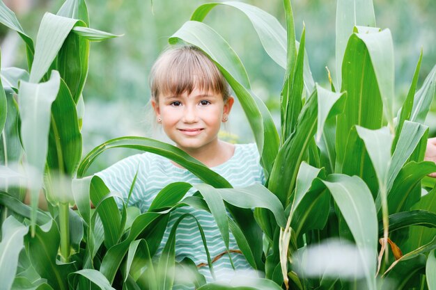 Lächelndes glückliches junges Mädchen und grüne Pflanzenblätter. Schöne niedliche kleine Frau, die auf Bauernhof im sonnigen warmen Tag steht. Attraktive Person und hohes Anbaukultur-Bio-Naturprodukt