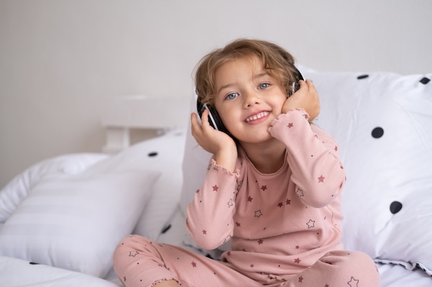 Lächelndes glückliches kleines Mädchen im Pyjama mit drahtlosen Kopfhörern, die zu Hause Musik im Bett hören.