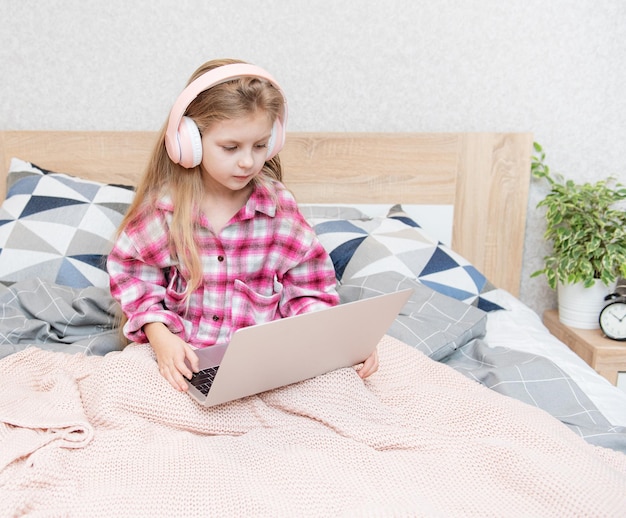 Lächelndes glückliches kleines Mädchen mit Laptop in drahtlosen Kopfhörern im Bett zu Hause.