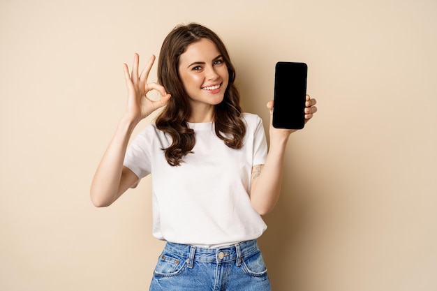 Lächelndes, glückliches Mädchen, das die Handy-Bildschirm-App auf dem Smartphone zeigt und in Ordnung ist, den Online-Shop zu empfehlen ...