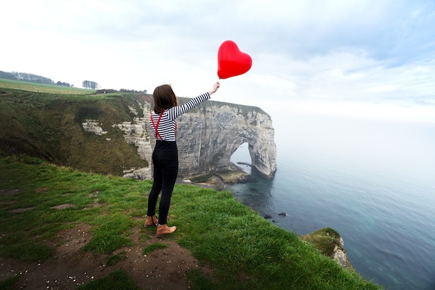 Lächelndes glückliches Mädchen mit einem roten Ballon in Form eines Herzens im Hintergrund der Landschaft Etretat. Frankreich
