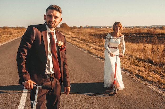 Lächelndes Hochzeitspaar, das bei Sonnenuntergang auf Rollern entlang der Straße außerhalb der Stadt fährt