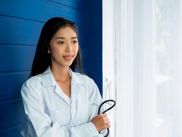 Lächelndes hübsches asiatisches Ärztinporträt, das auf blauem Holzhintergrund in der Nähe des Vorhangfensters in der Arztpraxis mit Kopierraum steht Selbstbewusste asiatische junge Ärztin mit Stethoskop