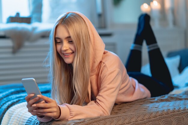 Lächelndes hübsches blondes Mädchen im rosa Parka und in den schwarzen Leggings unter Verwendung eines Smartphones, das zu Hause auf dem Bett liegt, während sie mit ihren Freunden plaudert