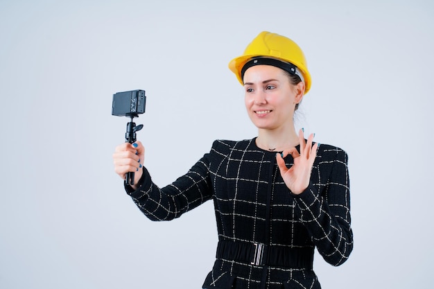 Lächelndes Ingenieurmädchen macht Selfie mit ihrer Kamera, indem sie perfekte Geste auf weißem Hintergrund zeigt