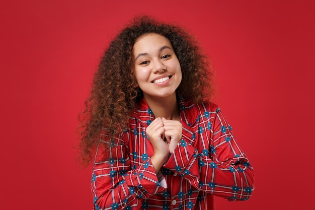 Lächelndes junges afrikanisch-amerikanisches Mädchen in Pyjama-Homewear, das posiert, während es sich zu Hause ausruht, isoliert auf rotem Hintergrund Studioportrait. Entspannen Sie sich bei guter Laune Lifestyle-Konzept. Mock-up-Kopienbereich. Zusammenballende Fäuste.