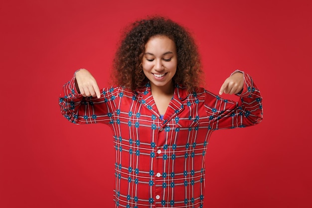 Lächelndes junges afrikanisch-amerikanisches Mädchen in Pyjama-Homewear, das posiert, während es sich zu Hause isoliert auf rotem Hintergrund ausruht. Entspannen Sie sich bei guter Laune Lifestyle-Konzept. Mock-up-Kopienbereich. Zeigefinger nach unten zeigen.