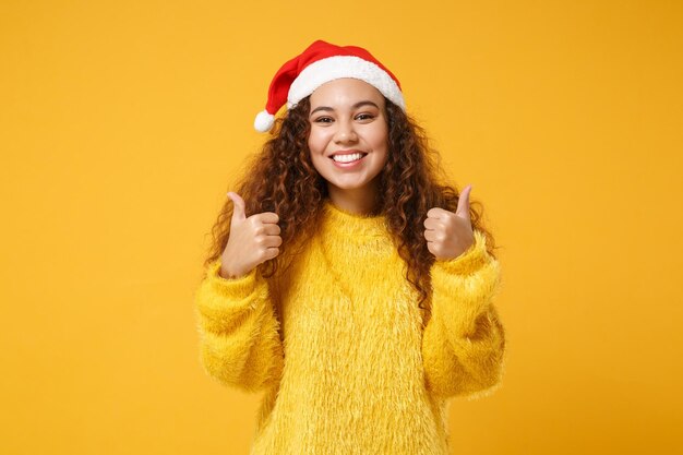 Lächelndes junges afroamerikanisches Weihnachtsmädchen im Pelzpullover, Weihnachtsmütze isoliert auf gelbem Hintergrund im Studio. Frohes neues Jahr 2020 Festtagskonzept. Mock-up-Kopienbereich. Daumen hoch zeigen.