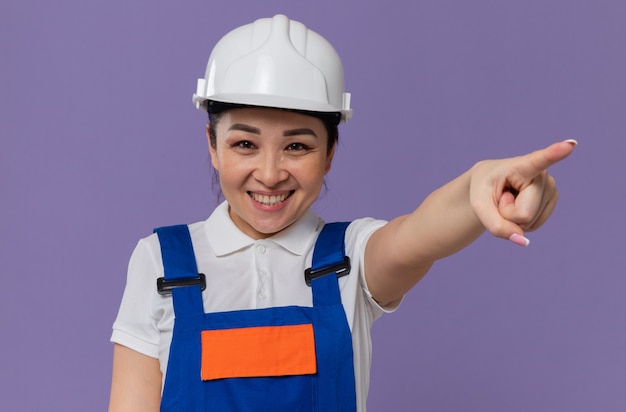 Lächelndes junges asiatisches Baumeistermädchen mit weißem Schutzhelm, der nach vorne schaut und auf die Seite zeigt