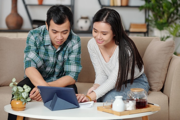 Lächelndes junges asiatisches Paar, das Tee trinkt und ein Video auswählt, um es zu Hause auf dem Tablet-Computer anzusehen?