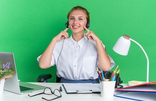 Lächelndes junges blondes Callcenter-Mädchen mit Headset am Schreibtisch sitzend mit Arbeitswerkzeugen, die mit den Fingern auf die Wangen zeigen