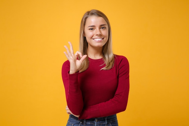 Lächelndes junges blondes Frauenmädchen in der beiläufigen Kleidung, die lokalisiert auf gelb-orangeem Wandhintergrund-Studioporträt aufwirft. Menschen aufrichtige Emotionen Lifestyle-Konzept. Mock-up-Kopienbereich. OK-Geste wird angezeigt.