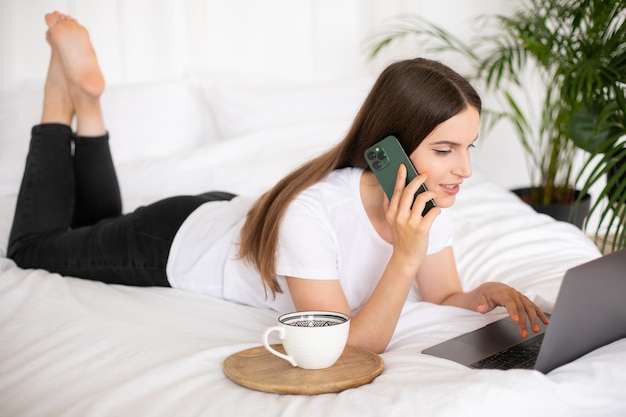 Lächelndes junges brünettes Mädchen, das zu Hause auf dem Bett liegt, an einem Laptop arbeitet und telefoniert