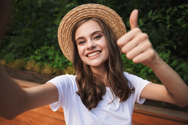 Lächelndes junges Mädchen, das ein Selfie mit ausgestreckter Hand im Park draußen nimmt und Daumen hoch zeigt