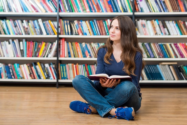 Lächelndes junges Mädchen, das mit einem offenen Buch auf den Knien auf dem Boden in der Bibliothek sitzt und zur Seite schaut, träumt oder sich etwas verschwommenes vorstellt Bücher im Hintergrund ein Konzept der Liebe zu Büchern