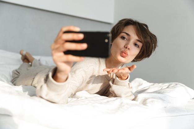 Lächelndes junges Mädchen, das sich morgens im Bett entspannt und ein Selfie macht