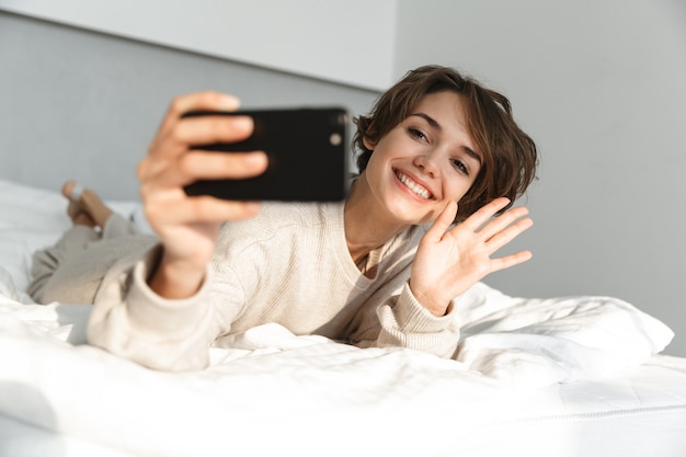 Lächelndes junges Mädchen, das sich morgens im Bett entspannt und ein Selfie macht